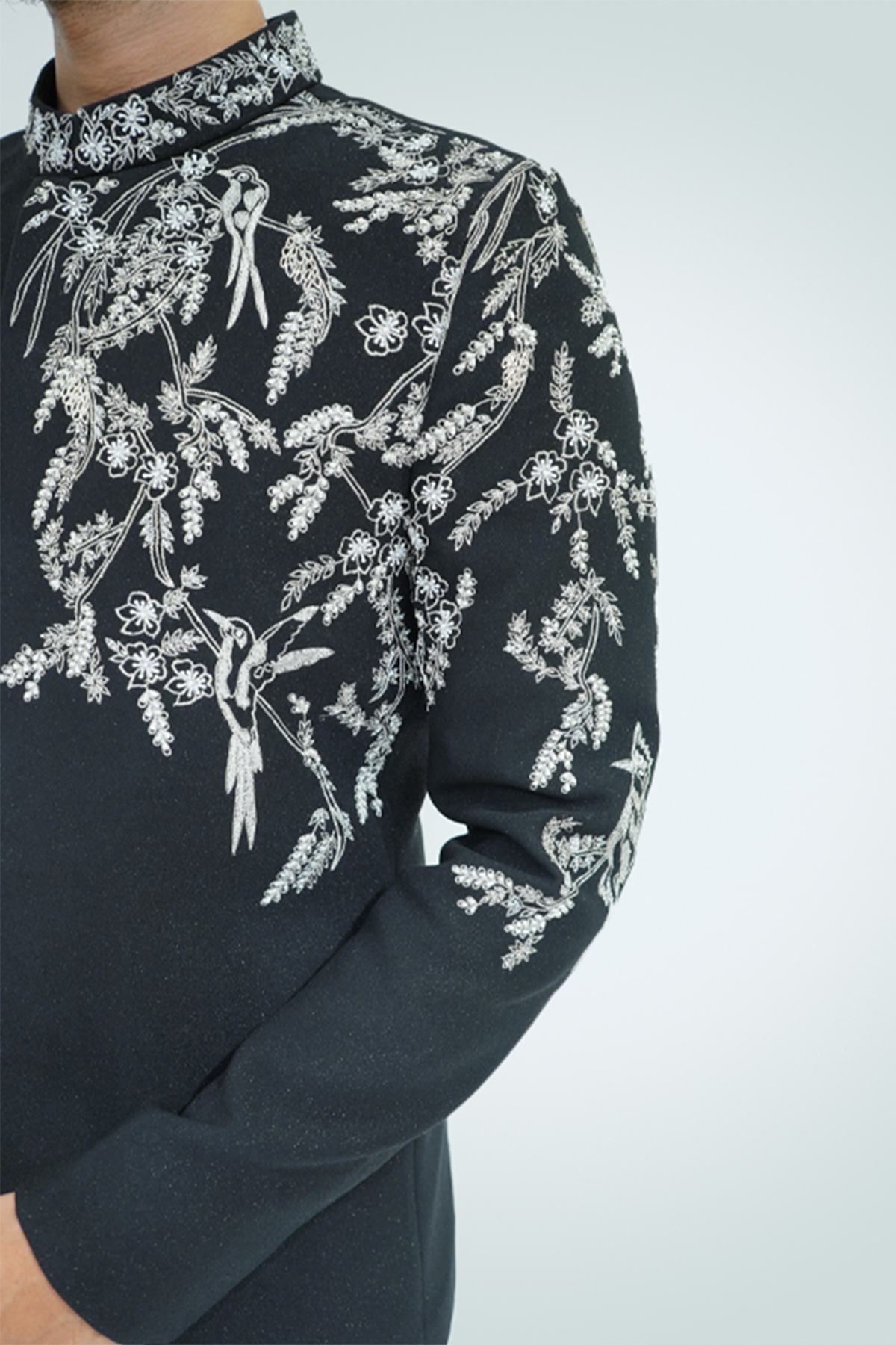 A man wearing a black suit with silver embroidery standing against a light background.
