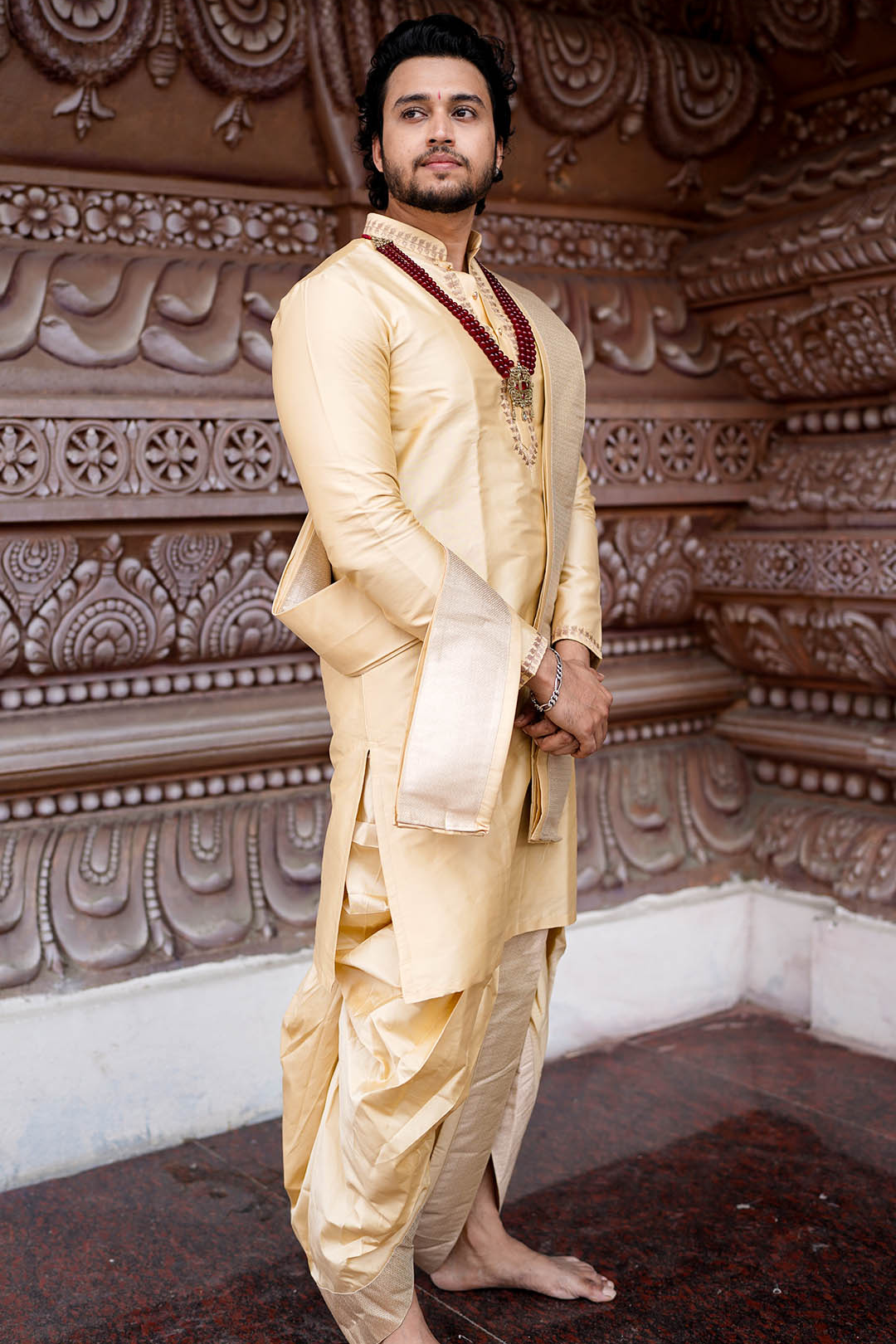 Man in a cream traditional outfit standing against an ornate wall.