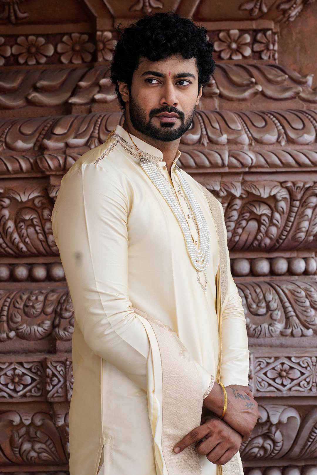 Man in traditional attire standing against a decorative wall.