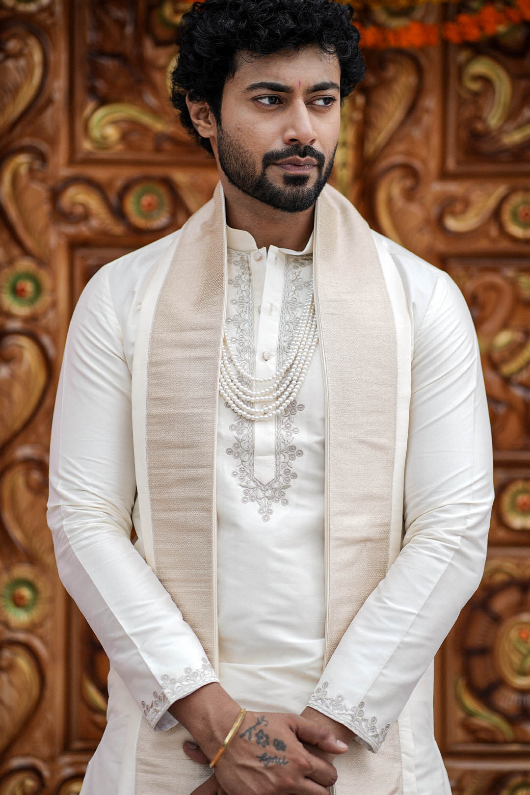 Man wearing a traditional white sherwani with intricate embroidery against a decorative wooden background