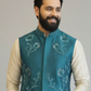 Man wearing a teal embroidered vest over a beige shirt on a gray background