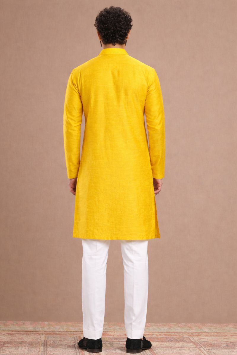 Person wearing a yellow kurta with white pants against a brown background
