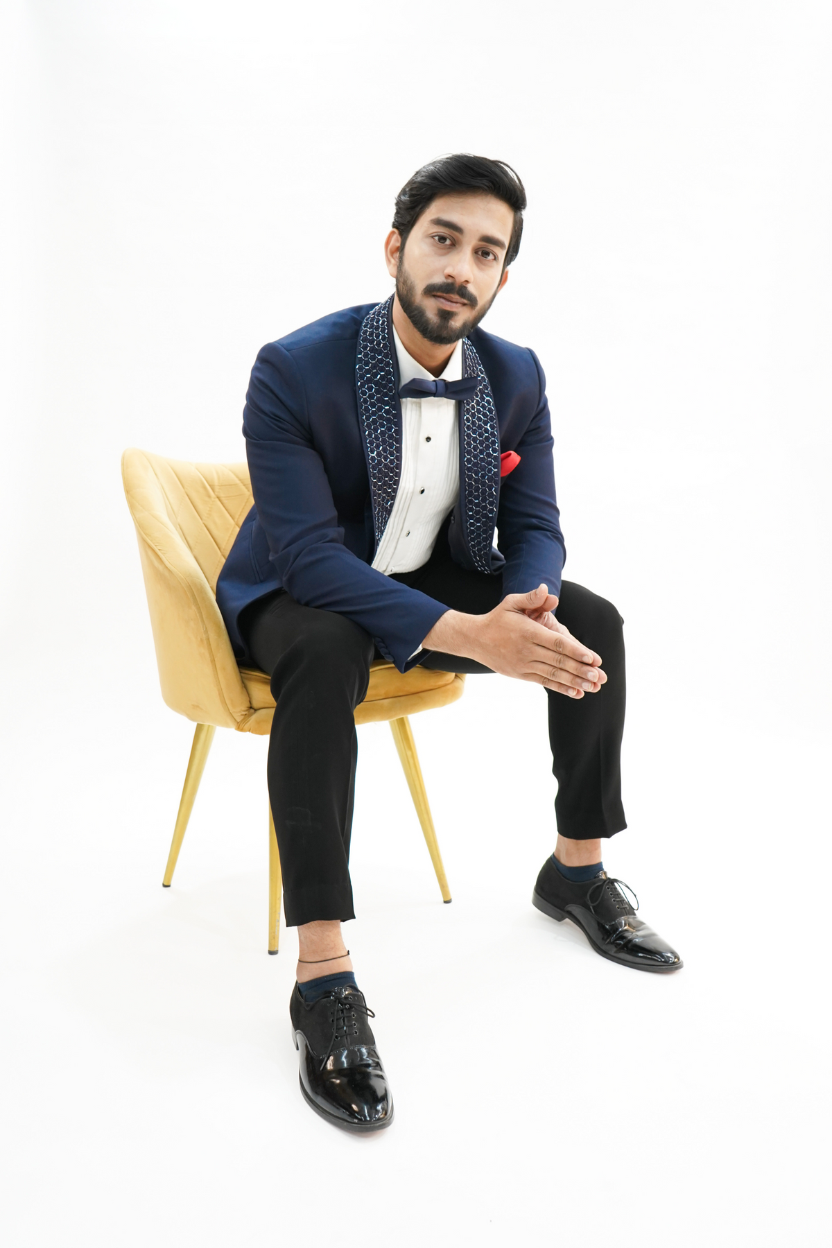 A man sitting on a yellow chair wearing a navy blue wool crepe 2-piece tuxedo with a detachable lapel, consisting of a jacket and trousers, accessorized with a bow tie and a handkerchief.