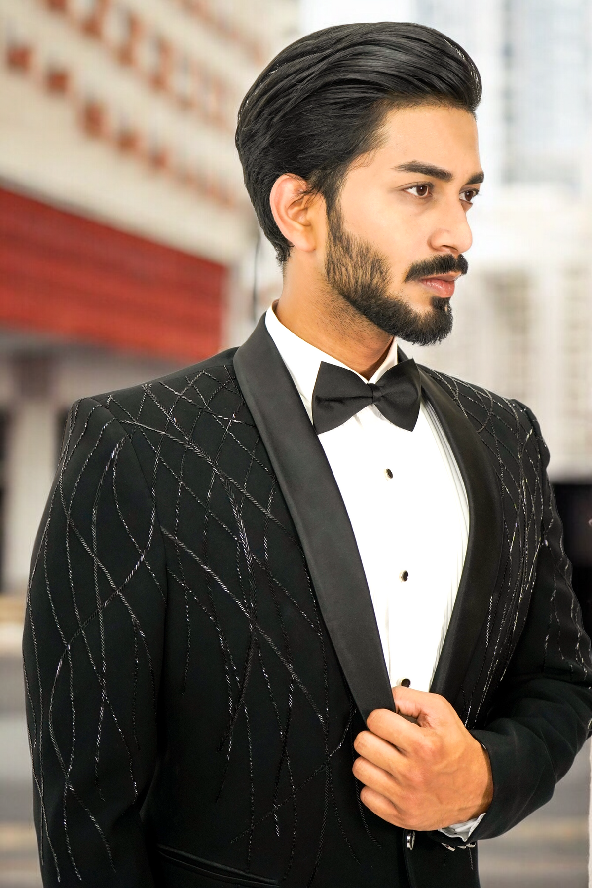 Man wearing a black tuxedo with a white shirt and bow tie, standing against a blurred indoor background.