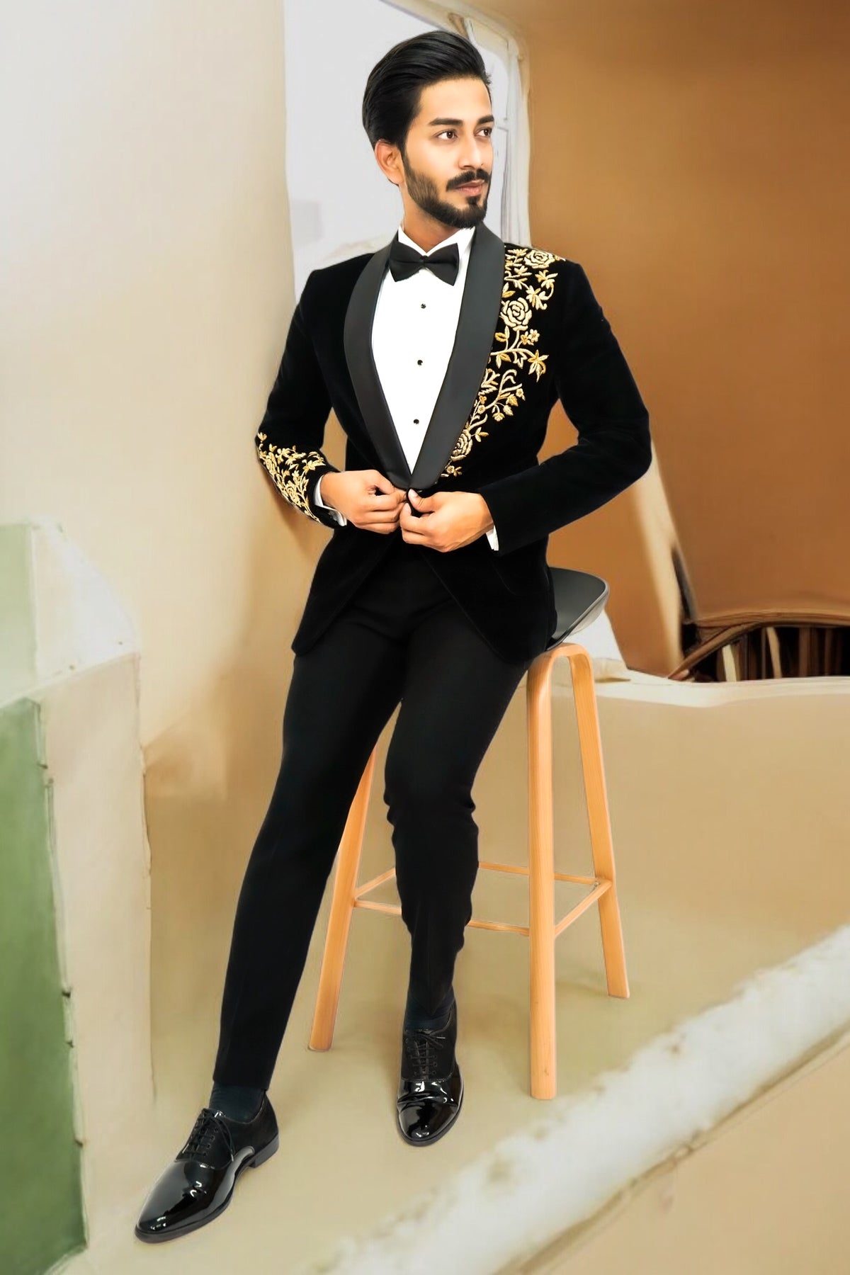 Man in a black tuxedo with gold embroidery sitting on a stool.