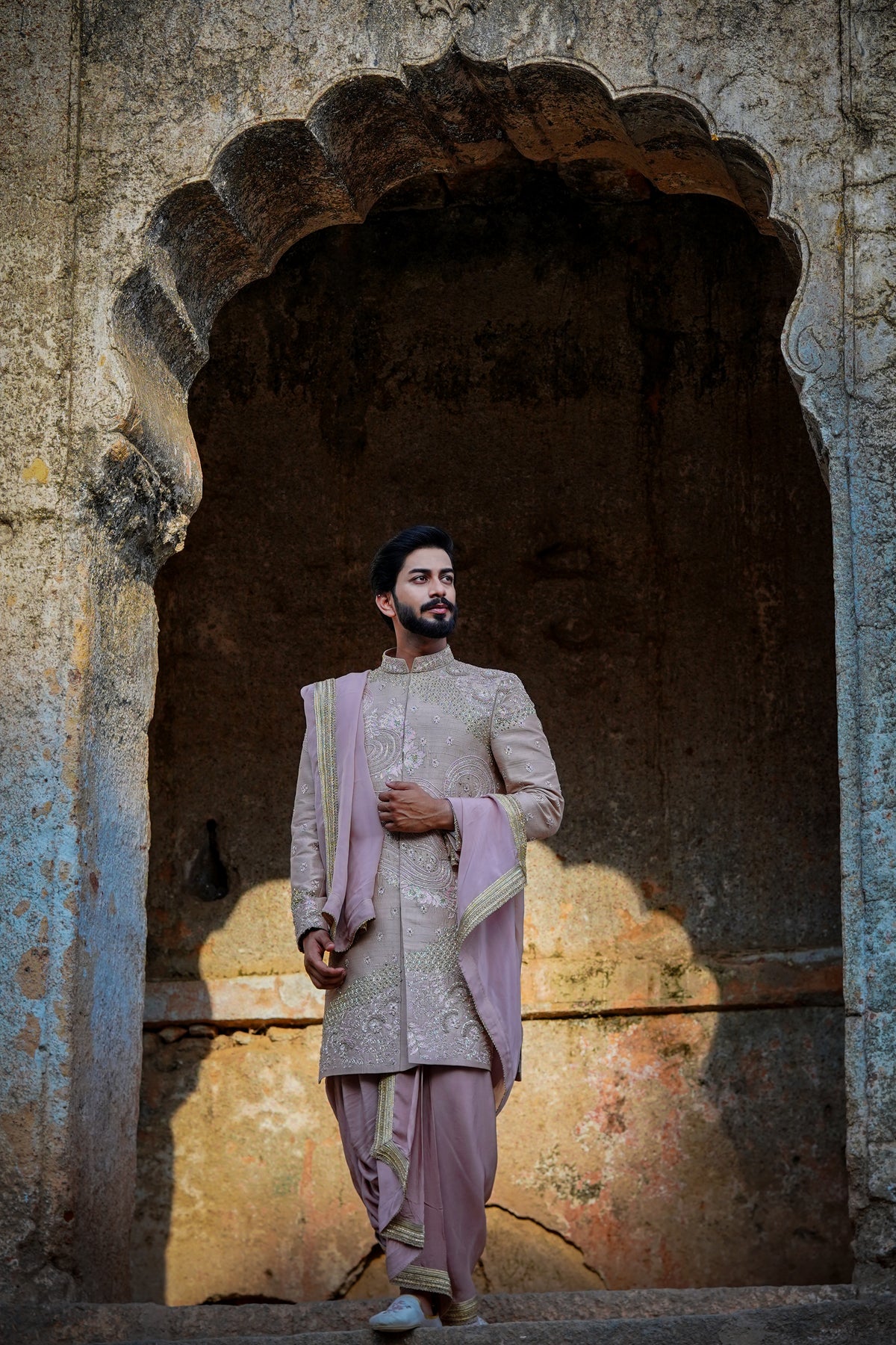A man wearing a traditional Sherwani with premium silk dhoti and khandwa, featuring intricate embroidery and a pink tint.