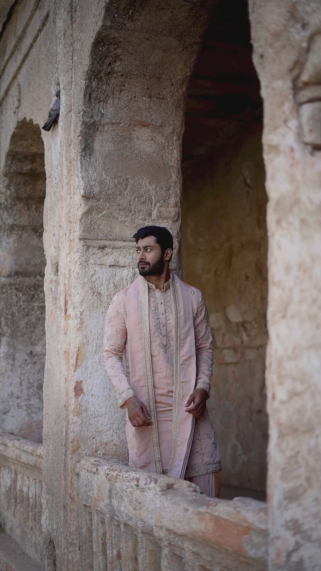 A man standing by a water body, wearing a traditional Sherwani with blue and gold colors, featuring a dupion silk dhoti and khandwa.