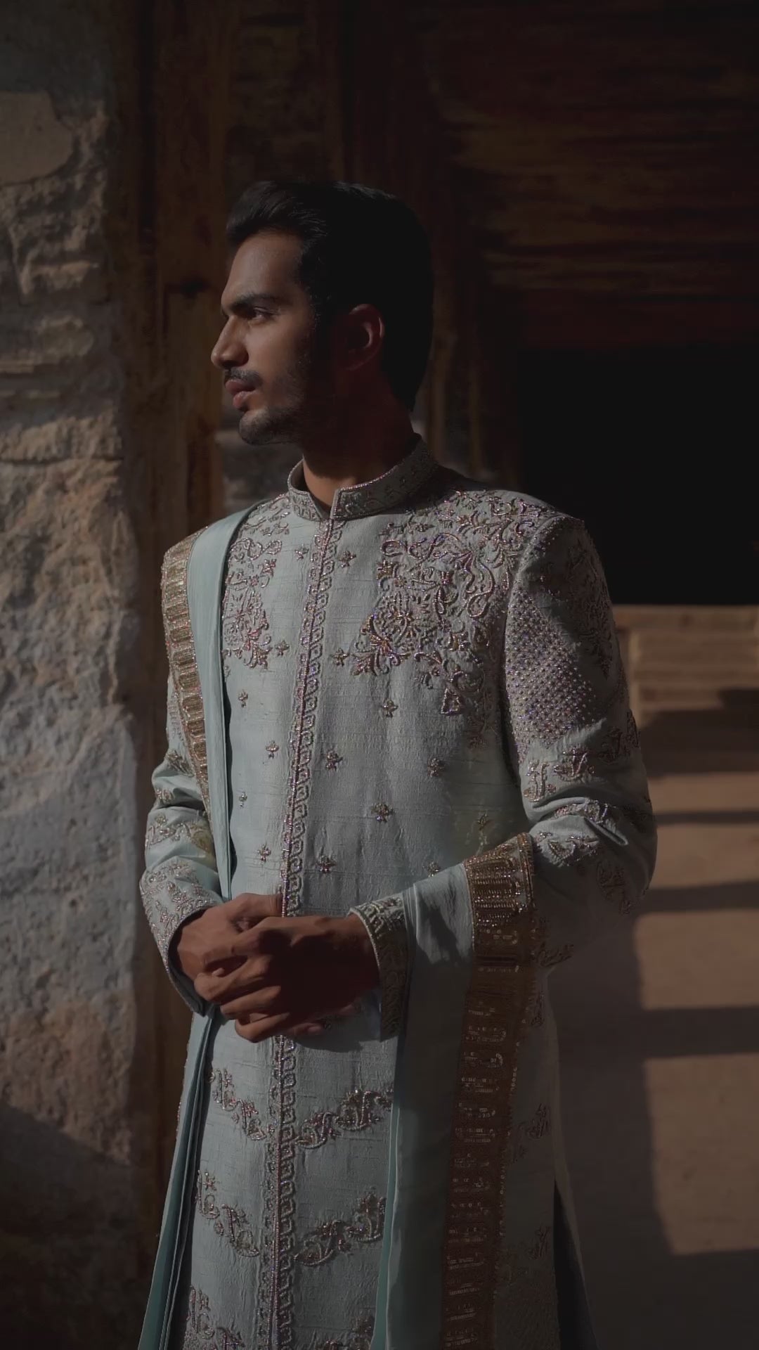 A man standing by a water body, wearing a traditional Sherwani with blue and gold colors, featuring a dupion silk dhoti and khandwa.