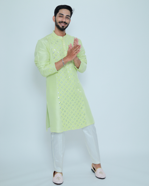 A man wearing a light green kurta with a mirror work embroidered panel, paired with white trousers and pink footwear.