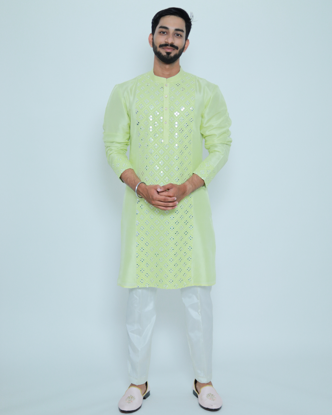 A man wearing a light green kurta with a mirror work embroidered panel, paired with white trousers and pink footwear.
