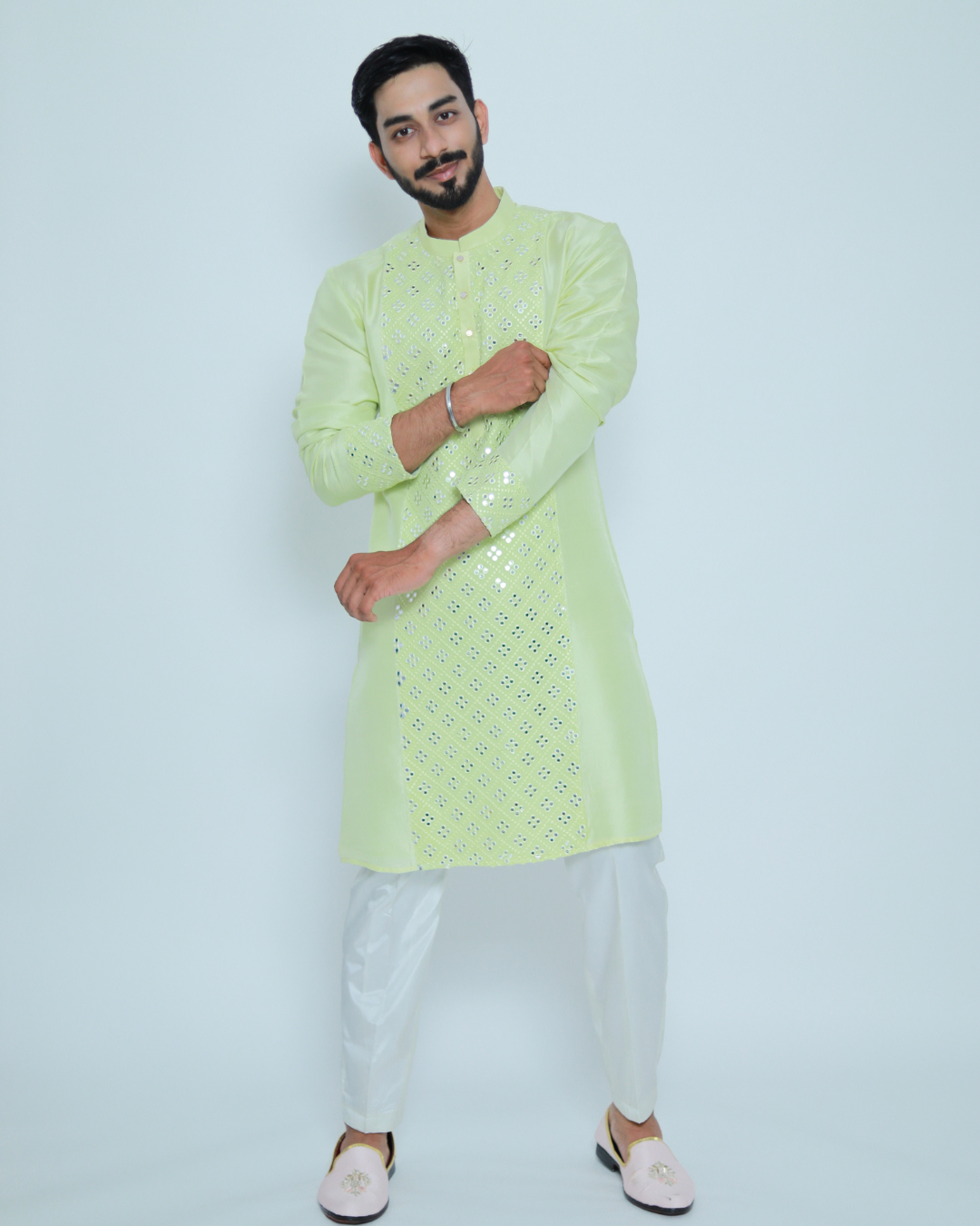 A man wearing a light green kurta with a mirror work embroidered panel, paired with white trousers and pink footwear.